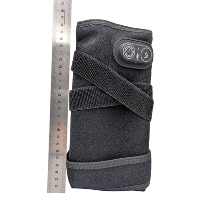 A dark gray fabric wrist wrap or arm sleeve with an adjustable hook-and-loop strap and a black control panel featuring two buttons, shown next to a metal ruler for scale on a white background.