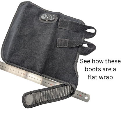 A dark gray heated boot wrap with an integrated control panel and straps, shown alongside metal rulers to demonstrate its flat wrap size, with text reading 'See how these boots are a flat wrap'.